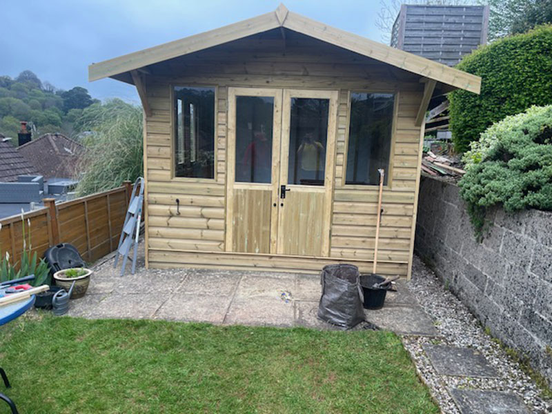 A wooden garden shed with double doors and windows, located at the edge of a grassy area. It's surrounded by a stone wall, plants, and gardening tools. The sky is overcast.