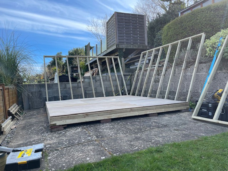 A partially constructed wooden structure is set on a paved area in a garden. Several wooden frames are propped up against a concrete wall. A grass lawn is in the foreground, and a clear sky is overhead.