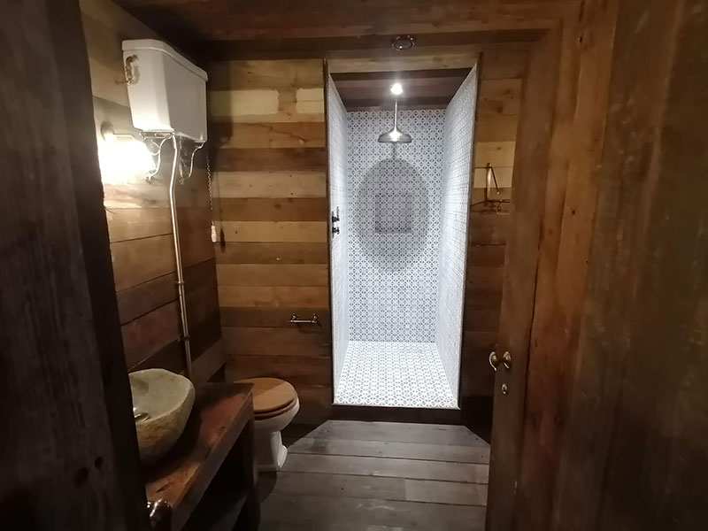 Wooden bathroom with rustic design, featuring a stone sink on a wooden countertop, a toilet, and a tiled shower area with a rain shower head. Warm lighting enhances the cozy atmosphere.
