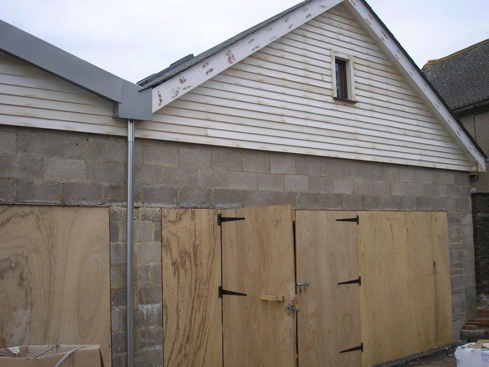 A building with a light-colored gabled roof and textured walls. The lower section of the walls features wooden boards with metal hinges, suggesting a garage or storage area. A downspout runs alongside the structure.
